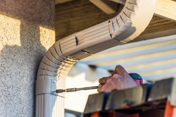 Worker Attaching Aluminum Rain Gutter and Down Spout to Fascia of House.